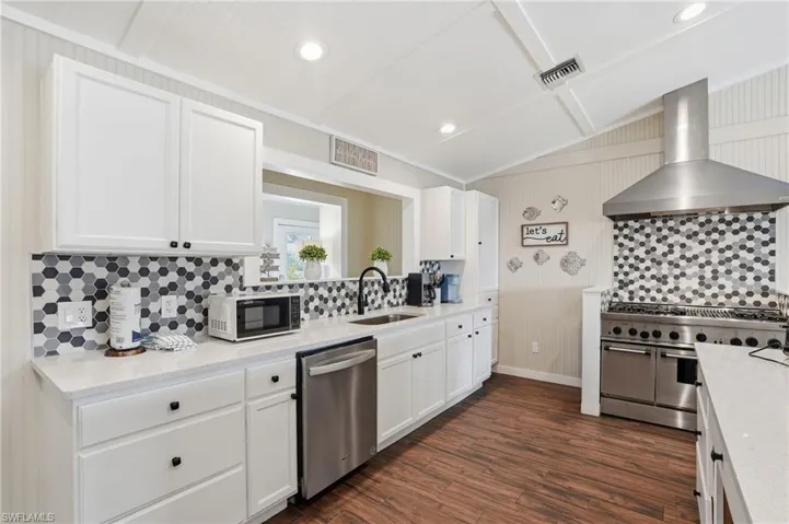 Kitchen featuring tasteful backsplash, white cabinetry, wall chimney range hood, appliances with stainless steel finishes, and lofted ceiling - Virtually Edited Image