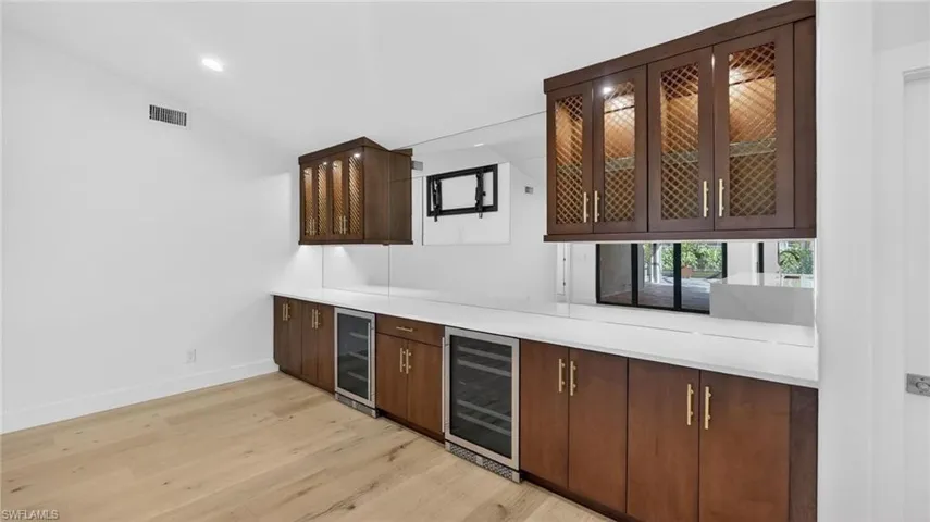Indoor bar with vaulted ceiling, wine cooler, light wood-style flooring, dark wood finish cabinetry, and glass fronted cabinets