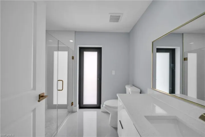 Modern bathroom featuring a frameless glass shower enclosure, a dual-sink vanity with a light-toned countertop, a rectangular mirror with a brass-finish frame, and white tile flooring