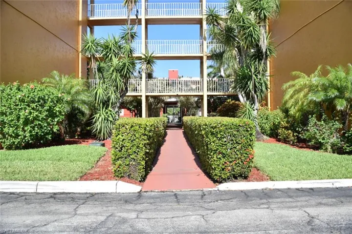 Exterior building facade with multiple balconies, featuring a red walkway bordered by manicured hedges and lush landscaping