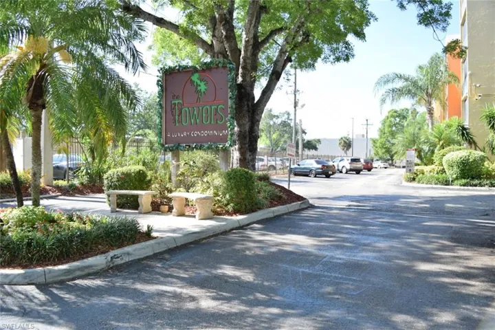 Entryway featuring a prominent community sign, stone benches, lush landscaping, mature trees, and palm trees