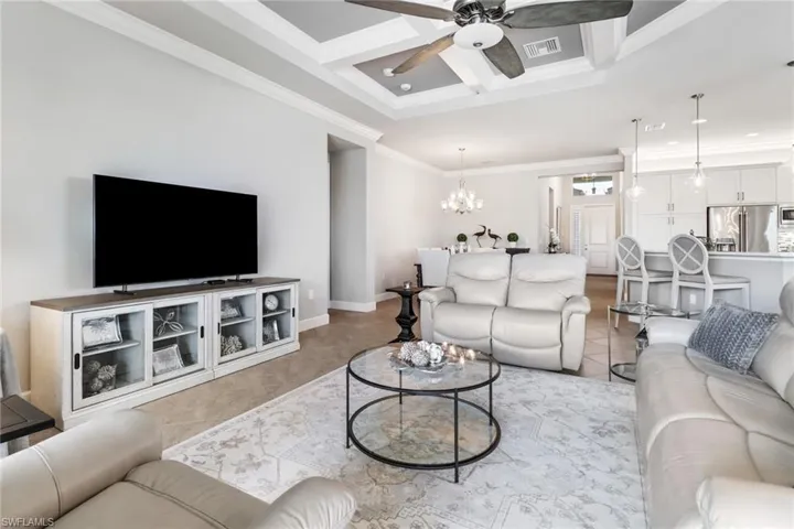 Living room with ornamental molding, coffered ceiling, ceiling fan with notable chandelier, and light tile patterned floors