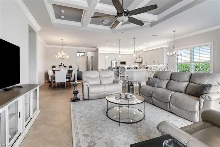 Living room featuring ceiling fan with notable chandelier, light tile patterned floors, ornamental molding, and coffered ceiling