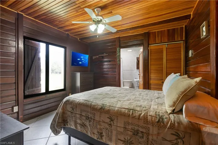 Bedroom with wooden walls, wood ceiling, light tile patterned flooring, a ceiling fan, and connected bathroom