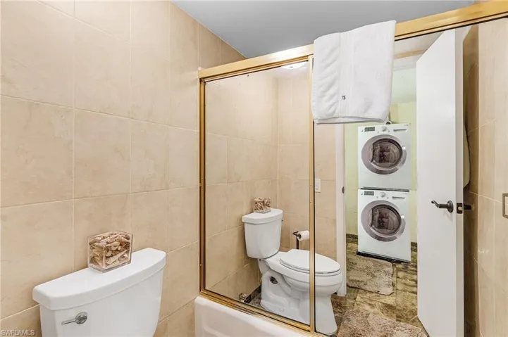 Full bath with tile walls, toilet, stacked washer / dryer, and enclosed tub / shower combo