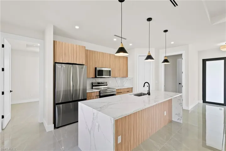 Kitchen featuring stainless steel appliances, light stone countertops, hanging light fixtures, a center island with sink, and light wood finish cabinets
