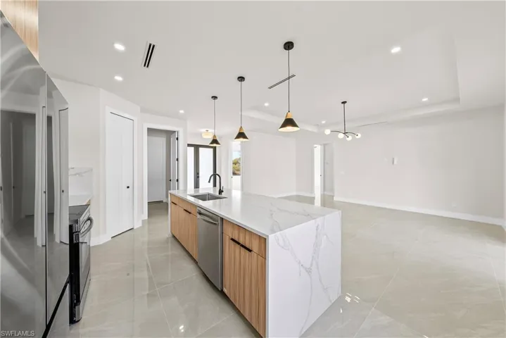 Kitchen featuring stainless steel appliances, light stone countertops, a tray ceiling, pendant lighting, and an island with sink