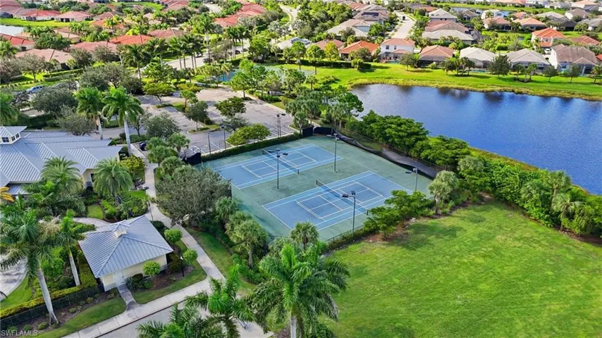 Recreational facilities featuring multiple tennis courts, a large green space, and a clubhouse with a metal roof, bordered by a substantial water body