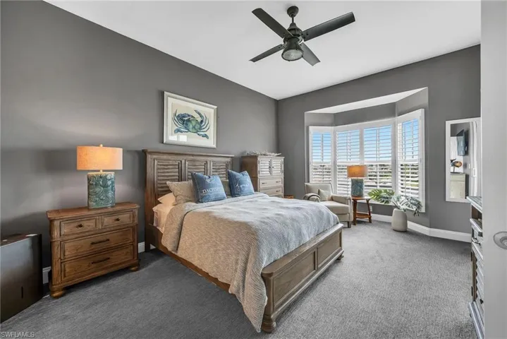 Spacious room featuring a bay window with plantation shutters, ceiling fan, and plush gray carpeting