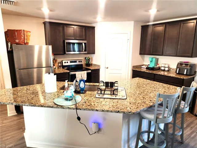 Kitchen featuring dark brown cabinets, stainless steel appliances, light stone counters, and an island with sink