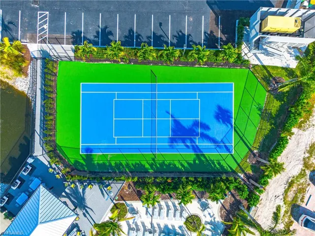 Regulation-size tennis court with blue playing surface and green surrounds, bordered by lush landscaping, a paved parking area, and a body of water