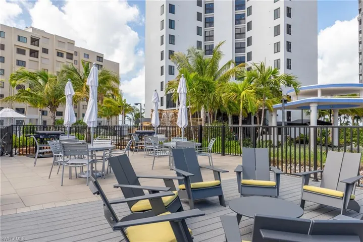 Resort-style poolside terrace featuring multiple lounge chairs with cushioned seating, white market umbrellas for shade, composite decking, black metal railings, and mature palm trees