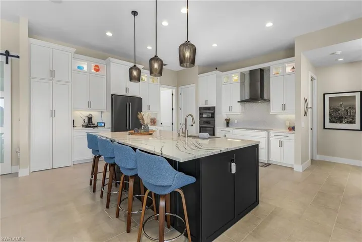 Kitchen with wall chimney exhaust hood, white cabinetry, a barn door, a kitchen island with sink, and black appliances