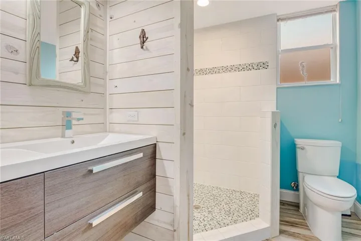 Floating wood-finish vanity with a rectangular sink, whitewashed horizontal paneled walls, shower with mosaic stone flooring and decorative tile inlay, and a frosted window