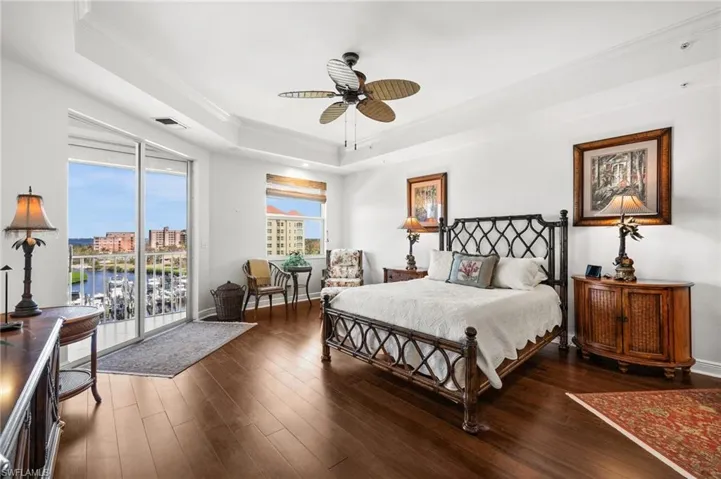 Primary bedroom featuring warm hardwood floors, a direct door to the screened lanai overlooking the marina. Notice the ceiling fan, tray ceilings, and elegant decor.