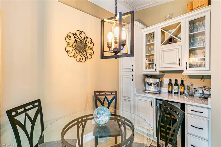 Indoor dry bar featuring a chandelier, beverage cooler, light stone countertops, white cabinetry, and glass insert cabinets - Virtually Edited Image