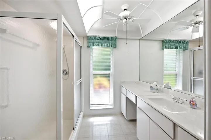 Bathroom featuring a shower enclosure with a sliding door, a dual-sink vanity with ample countertop space, a wall-mounted mirror, a ceiling fan, and a skylight