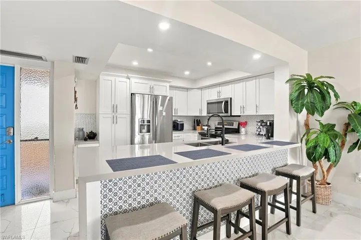 White kitchen featuring recessed lighting, a stone-finish countertop, a patterned tile island base, a stainless steel refrigerator, and a black faucet - Virtually Edited Image