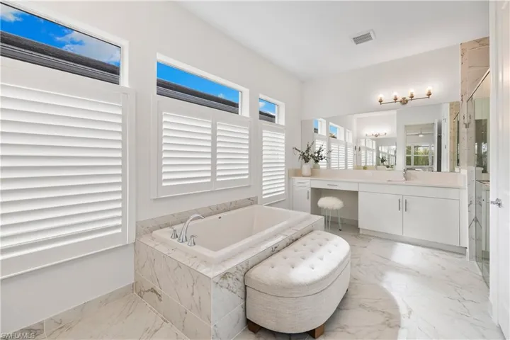 Spacious bathroom featuring a built-in soaking tub with tile surround, white plantation shutters, white vanity cabinetry, light-toned quartz countertops, and a glass-enclosed shower