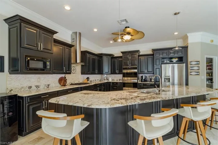 Kitchen featuring light stone counters, black appliances, wall chimney exhaust hood, a center island with sink, and tasteful backsplash