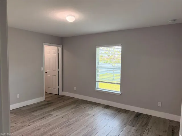 Interior room featuring wood-finish flooring, neutral wall paint, white baseboards, a single window with blinds, and a paneled door