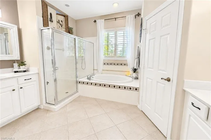 Bathroom featuring a built-in soaking tub with decorative tile surround, a separate glass-enclosed shower, dual vanity areas with white cabinetry and light-toned countertops, and light-colored tile flooring