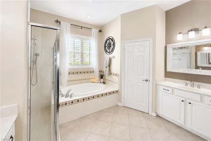 Bathroom featuring a built-in soaking tub with decorative tile surround, a glass-enclosed shower, dual vanity with white cabinetry, light-toned countertop, and tile flooring