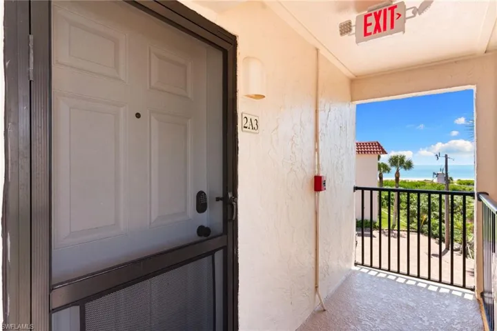Entry door with panel details and screen, textured exterior wall, exterior lighting fixture, black metal balcony railing, and ocean visible in the distance