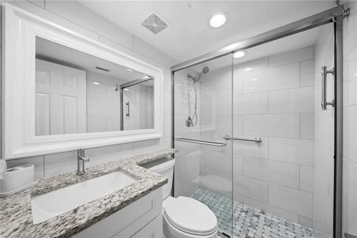 Granite-finish countertop, white shaker vanity, framed mirror, glass-enclosed shower, and mosaic tile shower floor