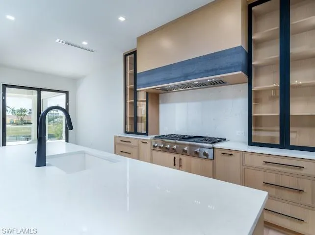 Kitchen with glass fronted cabinets, light wood finish cabinets, light stone counters, stainless steel gas stovetop, and recessed lighting