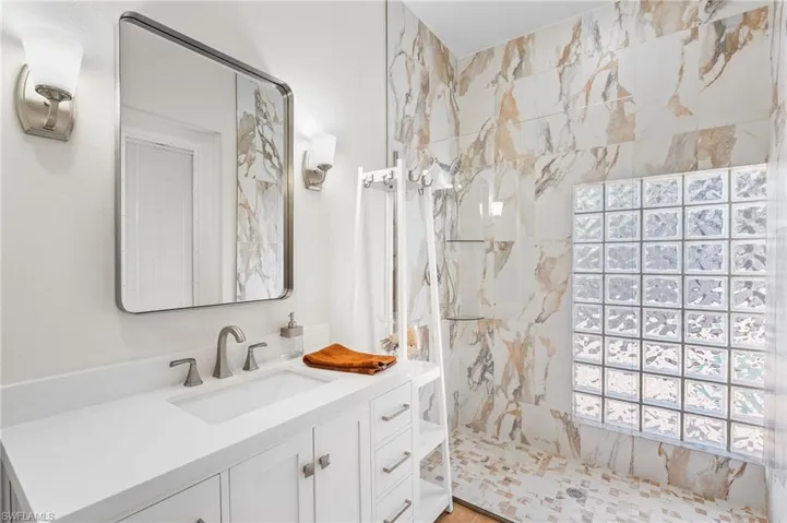 The second full bath is ideally designed as a dedicated pool bath, allowing effortless convenience without ever tracking through the home in wet attire. Just steps from the exterior entry, you are welcomed into a stunning walk-in shower adorned with floor-to-ceiling porcelain tile, paired with a sleek single vanity topped in quartz. An expansive glass block opening floods the space with natural light, enhancing the spa-like ambiance and refined coastal elegance.