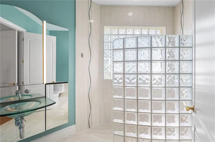 The third full bathroom, a beautifully appointed ensuite, offers a striking sense of refinement with a dramatic glass block feature wall that creates a seamless, walk-in shower experience. Floor-to-ceiling porcelain tile, accented with elegant listello detailing, adds a sophisticated architectural finish, while an expansive glass block window fills the space with radiant natural light. A sleek single glass vanity completes the design, preserving a clean, modern aesthetic with understated luxury.