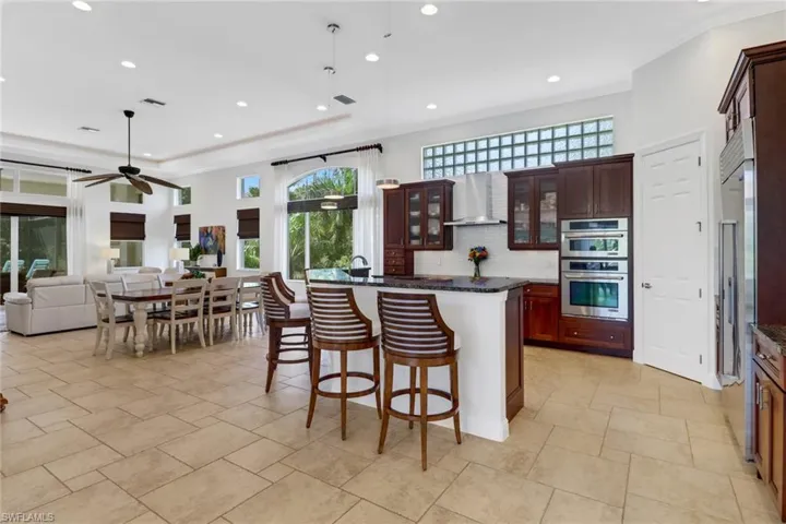 Kitchen with wall chimney range hood, an island with sink, backsplash, dark stone counters, and double oven