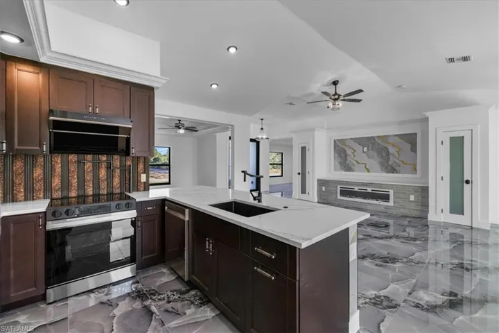 Kitchen with appliances with stainless steel finishes, light marble finish flooring, a glass covered fireplace, dark brown cabinets, and recessed lighting