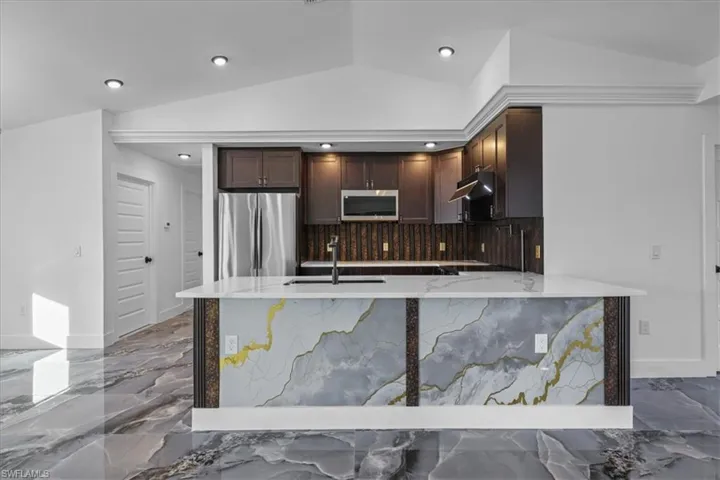 Kitchen with dark brown cabinetry, light stone counters, vaulted ceiling, appliances with stainless steel finishes, and dark marble finish floors