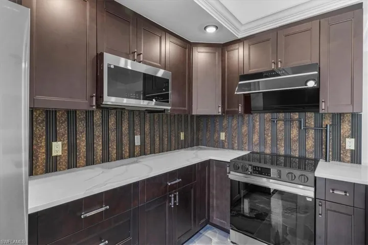Kitchen with dark brown cabinetry, appliances with stainless steel finishes, light stone counters, and ventilation hood