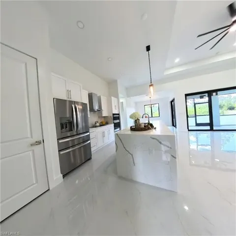 Kitchen featuring stainless steel fridge, wall chimney range hood, white cabinets, decorative light fixtures, and light stone countertops