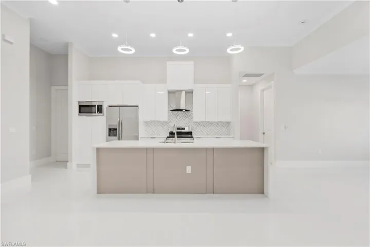 Kitchen featuring wall chimney exhaust hood, a center island with sink, appliances with stainless steel finishes, pendant lighting, and white cabinets