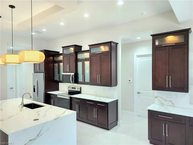 Kitchen featuring glass insert cabinets, light stone countertops, appliances with stainless steel finishes, hanging light fixtures, and dark brown cabinetry