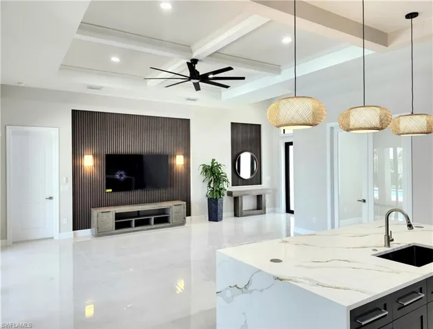 Kitchen with light stone countertops, an island with sink, hanging light fixtures, a ceiling fan, and beamed ceiling