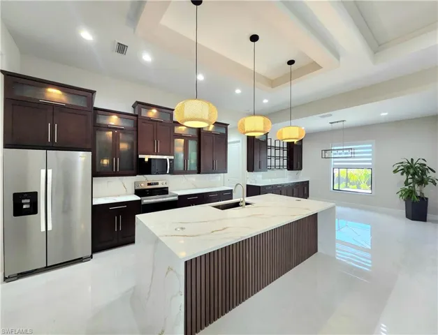 Kitchen featuring appliances with stainless steel finishes, a raised ceiling, dark brown cabinetry, glass insert cabinets, and light stone counters