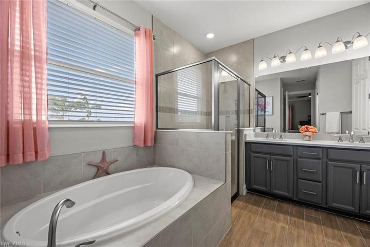 Luxurious primary bath with WOOD-LOOK TILE FLOOR, soaking tub, window blinds, double sink vanity with drawer stack, QUARTZ COUNTER, large shower with tile to the ceiling and decorative tile detail, and water closet.