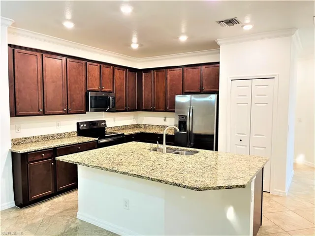 Kitchen with stainless steel appliances, light stone counters, dark wood finish cabinets, a center island with sink, and ornamental molding
