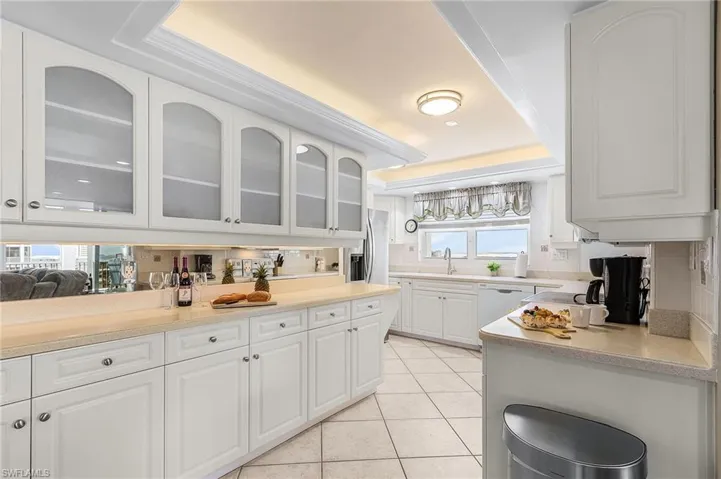 Kitchen featuring white cabinetry, light tile patterned flooring, a raised ceiling, glass insert cabinets, and stainless steel fridge with ice dispenser - Virtually Edited Image