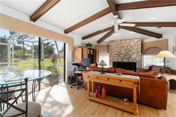 Living room with a stone fireplace, ceiling fan, lofted ceiling with beams, and light wood-type flooring