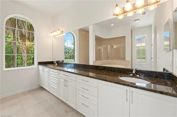 Bathroom featuring a dual vanity with a dark countertop and white cabinetry, arched windows, light tile flooring, a large wall mirror, and a built-in tub with a tiled surround