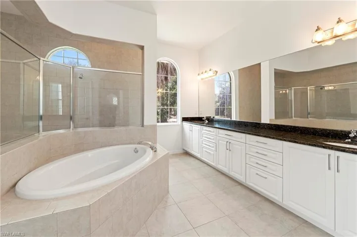 Built-in soaking tub within a tiled surround, featuring a glass-enclosed shower, dual arched windows, an extended vanity with dual sinks, and white shaker-style cabinetry