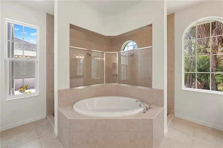 Corner soaking tub with a tiled surround, framed by a glass-enclosed shower featuring a tiled interior and an arched window
