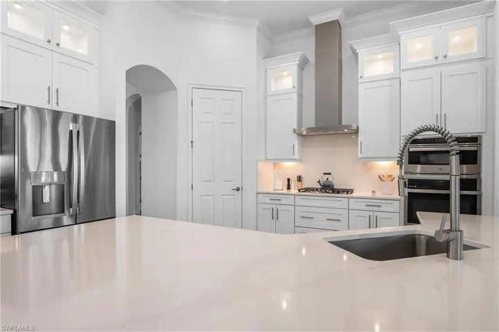 Kitchen featuring stainless steel appliances, glass insert cabinets, white cabinetry, ornamental molding, and wall chimney range hood