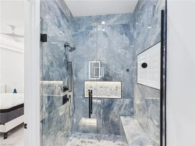 Glass-enclosed shower featuring blue-grey stone-look tile walls, matte black fixtures, a built-in bench, and an illuminated mosaic tile niche